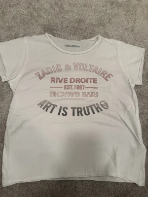 Vit Zadig & Voltaire t-shirt - Vit t-shirt från Zadig & Voltaire med tryck i rött och svart framtill. Texten 'RIVE DROITE', 'ART IS TRUTH' och en liten döskalle ger en cool vibe. Klassisk rund hals och korta ärmar. Perfekt till jeans eller shorts.