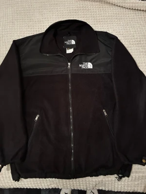 Svart fleecejacka från The North Face - Säljer en svart fleecejacka från The North Face med dragkedja framtill och två sidofickor med dragkedja. Jackan har detaljer i syntetmaterial över axlar och bröst samt broderad logga på bröstet. Perfekt för kyliga dagar och outdoor vibes. Tröjan är i storlek XXL men mer som XL