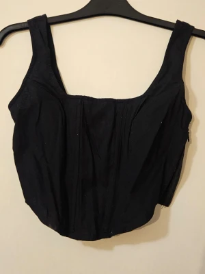 Svart korsett-topp med breda axelband - Stilren svart korsett-topp med breda axelband och markerade sömmar framtill. Toppen har en smockad rygg för extra komfort och en diskret dragkedja i sidan.