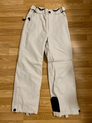 Vita täckbyxor med svarta detaljer - Snygga vita täckbyxor från CLWR med svarta detaljer och förstärkning vid benslut. Byxorna har justerbar midja, dragkedjor vid benslut och praktiska fickor. Strl 164 junior.