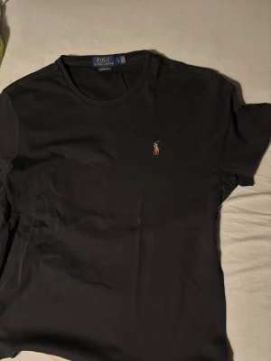 Svart t-shirt från Polo Ralph Lauren - Klassisk svart t-shirt från Polo Ralph Lauren med rund hals och broderad logga på bröstet. Modellen är custom slim fit och har korta ärmar. Perfekt basplagg med stilren design och diskret detalj.