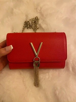 Röd axelväska från Valentino - Snygg röd axelväska från Valentino med silverfärgad kedja och V-detalj framtill. Väskan har en stilren, rektangulär form och är tillverkad i skinn med struktur. Kedjan och tofsen i metall ger en lyxig touch. Perfekt för att lyfta din outfit.