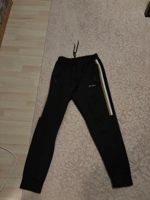 Svarta mjukisbyxor från Hugo boss - Säljer ett par svarta mjukisbyxor från Hugo boss med vita och bruna sidoremsor. Byxorna har dragkedjeförsedda fickor och resår i midjan med snörning. Perfekta för chill dagar eller träning. Materialet känns mjukt och bekvämt.