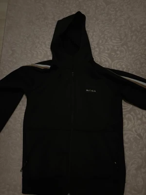 Svart hoodie från BOSS med dragkedja - Svart hoodie från BOSS med dragkedja framtill och huva. Snygga detaljer med vita och bruna ränder längs ärmarna samt BOSS-logga på bröstet. Perfekt för en stilren och sportig look. Tillverkad i mjukt och bekvämt material.