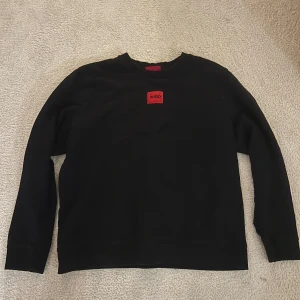 Svart sweatshirt från Hugo - Säljer en svart sweatshirt från Hugo med klassisk rund hals och en röd patch med logga på bröstet. Tröjan har en avslappnad passform och är tillverkad i mjukt material som passar perfekt till jeans eller joggers.