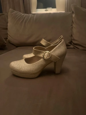 Glittriga guldiga pumps med rem - Snygga pumps i glittrigt guld med rund tå, hög klack och bred rem med dekorativt spänne. Skorna har en stadig plattform och ger en riktigt festlig look. Perfekta för dig som vill sticka ut med stil.