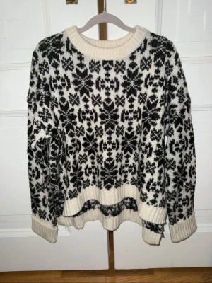 Svartvit stickad tröja från H&M  - Stickad tröja i svart och vitt från H&M med klassiskt nordiskt mönster. Tröjan har rund halsringning, ribbade muddar och nederkant. Perfekt för kalla dagar och ger en mysig vinterlook.