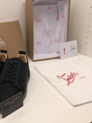 Svarta sneakers från Christian Louboutin - Snygga svarta sneakers från Christian Louboutin med klassisk röd sula och svarta skosnören. Skorna har en stilren design och är tillverkade i tyg med detaljer i skinn. Perfekta för dig som vill ha något exklusivt och trendigt.