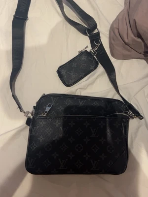 Svart axelväska  - Snygg svart axelväska med klassiskt monogrammönster och justerbar axelrem. Väskan har en framficka med dragkedja och en matchande liten plånbok som hänger i remmen. Perfekt för att bära dina viktigaste saker!