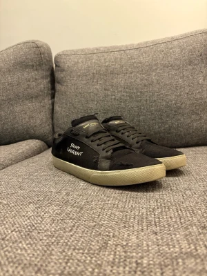 Svarta Saint Laurent sneakers - Svarta sneakers från Saint Laurent med vit text på sidan och klassisk låg siluett. Skorna är i väldigt bra skick! Skorna har snörning och klassisk vit/gul sula. Materialet är textil och skinn, vilket ger en stilren och urban look. !!OBS!! Slitningarna kommer med när man köper nya! Kan även gå ner i pris vid snabb affär