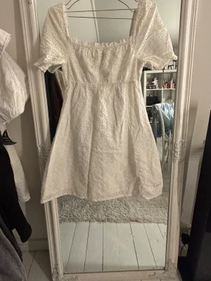 Vit kort klänning från GANT - Söt vit kort klänning från GANT med broderade detaljer och puffärmar. Klänningen har en fyrkantig ring sop neckline och är tillverkad i bomull, vilket gör den både luftig och bekväm. Perfekt för sommarens alla tillfällen!
