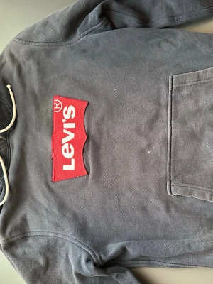 Mörkgrå Levi's hoodie med logga - Säljer en mörkgrå hoodie från Levi's med stor röd och vit logga på bröstet. Hoodien har huva med vita snören och en klassisk magficka. Tillverkad i mjuk bomullsblandning som är skön att ha på sig. Perfekt för en avslappnad och cool stil.