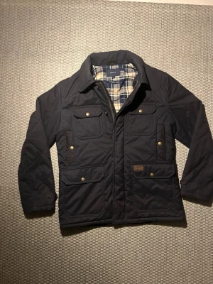 Mörkblå quiltad jacka från Gant - Snygg mörkgrå quiltad jacka från Gant med rutigt innerfoder. Jackan har fyra fickor med lock och tryckknappar, dragkedja och knappar framtill samt klassisk krage. Perfekt för kyligare dagar och ger en stilren look. Passar även L. Inga defekter