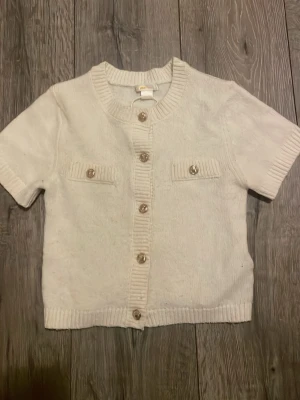 Vit kortärmad stickad kofta - Kortärmad vit stickad kofta med ribbade kanter och dekorativa knappar framtill. Två små fickor med knappdetaljer på bröstet ger en trendig touch. Perfekt att bära över ett linne eller t-shirt för en avslappnad men stilsäker look.