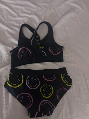 Svart bikini Smiley World x H&M - Säljer en svart bikini från Smiley World x H&M med färgglada smiley-mönster i rosa och gult. Överdelen har brottarrygg och bred resår, medan underdelen har klassisk passform. Materialet är stretchigt och bekvämt, perfekt för stranden eller poolen.