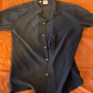 Mörkblå ribbad skjorta med knappar Zara  - Snygg mörkblå skjorta med ribbad struktur och klassisk krage. Skjortan har knappar framtill och korta ärmar, perfekt för en avslappnad men stilren look.