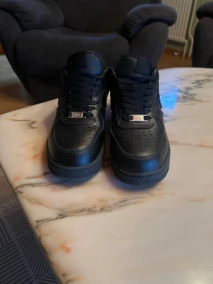 Svarta Nike Air Force 1 sneakers - Klassiska helsvarta Nike Air Force 1 sneakers med låg profil och rund tå. Skorna har snörning och är tillverkade i skinn med diskret Nike-logga på sidan. Perfekta för dig som gillar stilrena och tidlösa sneakers.