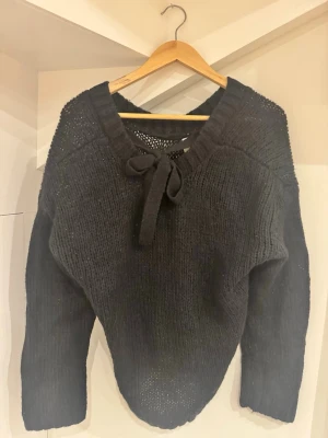 Svart stickad tröja med knyt från kay/day - Mysig svart stickad tröja från kay/day i storlek XS. Tröjan har en bred ribbad ringning med knytband framtill och långa ärmar. Perfekt för kyliga dagar och enkel att styla med jeans eller kjol.
