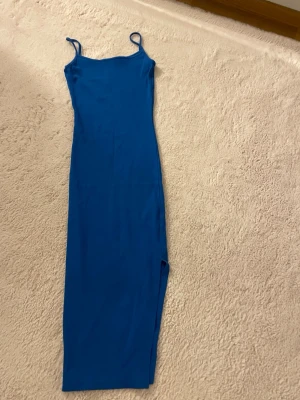 Blå långklänning med slits - Blå långklänning från New Yorker med smala axelband och snygg slits på sidan. Figurnära passform i stretchigt ribbat material. Perfekt för sommaren, stranddagar eller festliga tillfällen. Endast lätt använd.