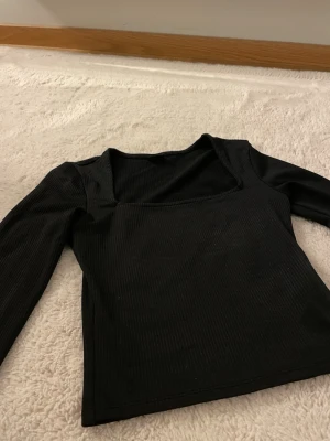 Svart ribbad långärmad topp - Trendig svart ribbad topp med lång ärm och fyrkantig ringning. Croppad design i stretchigt material som sitter bekvämt. Enkel men stilren, passar perfekt till både jeans, kjol eller under en kavaj.