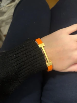 Hermesliknande armband - Säljer ett stilrent armband i orange läder med en stor guldfärgad metallspänne i form av ett H. Armbandet har en elegant och modern look, perfekt för att lyfta vilken outfit som helst. Justerbart spänne för enkel användning.