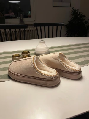 Beige fluffiga tofflor storlek 40-41 - Mysiga beige tofflor från HOME i storlek 40-41. Utsidan är i mjuk mockaimitation och insidan är fodrad med fluffigt vitt foder. Dekorativ söm i beige och gult runt öppningen och en rejäl platt sula i beige. Perfekta för kalla dagar hemma.