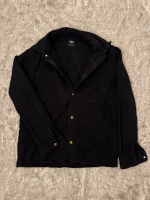 Svart jeans skjorta  - Svart jeans skjorta i storlek M från Zara med klassisk krage och tryckknappar framtill. Två bröstfickor och långa ärmar med knapp vid ärmslut. Kontakta gärna om ni har några frågor eller funderingar gällande pris eller plagg