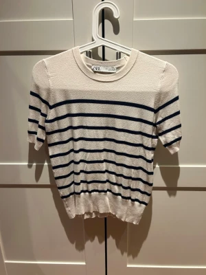 Randig topp från Zara i bomull - Kortärmad topp från Zara i mjuk bomull. Vit botten med mörkblå horisontella ränder, rund halsringning och ribbad kant nedtill. Perfekt till jeans eller kjol för en enkel och fräsch look.