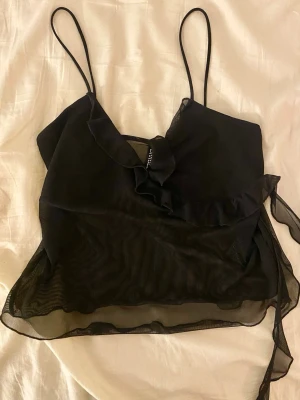Svart mesh omlottopp från H&M Divided - Säljer en svart mesh omlottopp från H&M Divided med tunna axelband och volangdetaljer framtill. Toppen är genomskinlig och har knyt i sidan för en snygg passform.