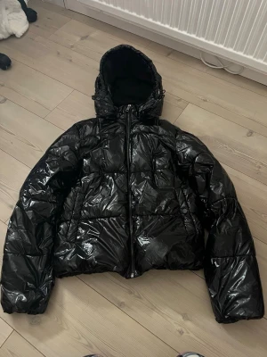 Svart pufferjacka från Nelly🩷 - Säljer en glansig svart pufferjacka från Nelly med huva och dragkedja framtill. Jackan har en oversized vibe och är perfekt för kalla dagar. Tillverkad i syntetmaterial och har två stora fickor framtill. Riktigt snygg och trendig look!🩷