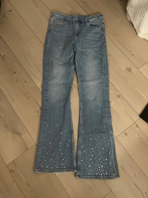 Coola blå lowwaist bootcut jeans Gina Tricot 164 - Säljer ett par ljusblå bootcut jeans från Gina Tricot i storlek 164. Jeansen har råa, fransiga benslut och är dekorerade med vita pärlor nertill på båda benen. Klassisk femficksmodell i mjukt denimtyg. Aldrig andvända. Perfekta för dig som vill ha något extra till din outfit!🩷