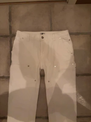 Vita bootcut byxor från Filippa K - Säljer ett par vita bootcut-byxor från Filippa K. Byxorna har raka ben med lätt utsvängning, silverfärgade nitar och knapp, samt praktiska fickor fram. Tillverkade i ett mjukt bomullsmaterial med klassisk midja och bälteshällor.