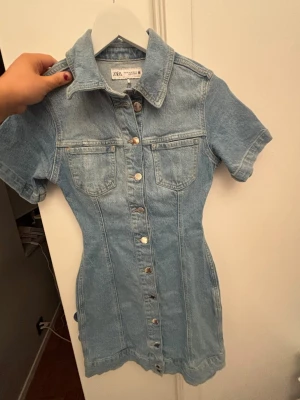 Jeansklänning från Zara - Säljer en ljusblå jeansklänning från Zara med korta ärmar, krage och knappar hela vägen framtill. Klänningen har två bröstfickor och markerad midja för en snygg siluett. Perfekt till sneakers eller boots för en cool look.