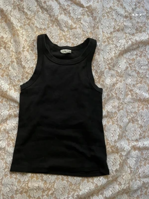 Svart ribbad linnetopp - Säljer en svart ribbad linnetopp med breda axelband och rundad halsringning. Toppen är figurnära och har en klassisk, enkel design som passar till mycket. Materialet känns mjukt och stretchigt, perfekt till jeans eller kjol.