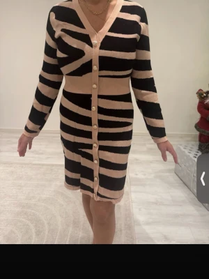 Beige och svart randig stickad klänning - Snygg kort stickad klänning i beige och svart med breda ränder och zebrainspirerat mönster. Klänningen har v-ringning, långa ärmar och knappar hela vägen framtill. Figurnära passform som markerar midjan och ger en cool look.