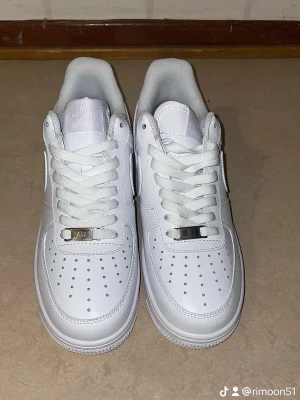 Nike Air Force 1 helvita sneakers - Klassiska Nike Air Force 1 sneakers i helvitt skinn med perforerad tå, rund tå och vit sula. Skorna har snörning med metallbricka och den ikoniska swoosh-loggan på sidan. Perfekta för dig som vill ha en clean och tidlös look.