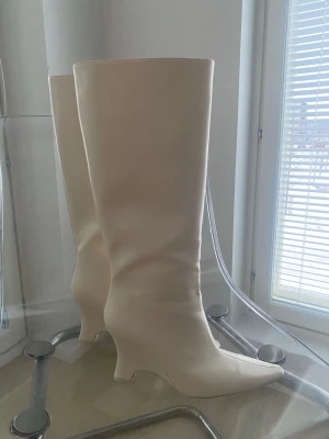 Beiga höga boots med spetsig tå - Säljer ett par stilrena höga boots i beige skinnimitation. Skorna har spetsig tå, låg klack och slät design utan synliga detaljer. Perfekta för dig som vill ha en clean och trendig look till höst och vinter.