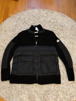 Moncler Cardigan  - En svart Moncler-jacka i hybridstil med stickade ärmar och rygg samt vadderad framsida. Hel dragkedja, två bröstfickor med knapp och klassisk Moncler-logga på ärmen.  Storlek XL men passar L, Skick 9/10.  Kvitto finns!!! Det är bara o skriva vid funderingar!