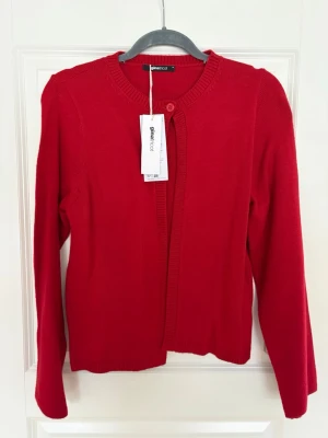 Röd kofta från Gina Tricot M oanvänd  - Klassisk röd kofta från Gina Tricot med rund halsringning och ribbade kanter. Långärmad modell med knapp framtill. Perfekt att slänga över en topp för en enkel men snygg look.