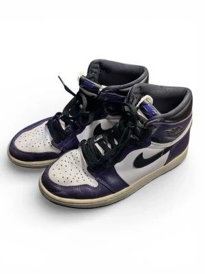 Nike Air Jordan 1 Court Purple - Säljer ett par Nike Air Jordan 1 Court Purple med svart swoosh, vita paneler och lila detaljer. Skorna har svarta skosnören, perforerad tåbox och klassisk hög siluett. Tillverkade i skinn med vadderad krage för extra komfort. Perfekta för dig som gillar streetwear.