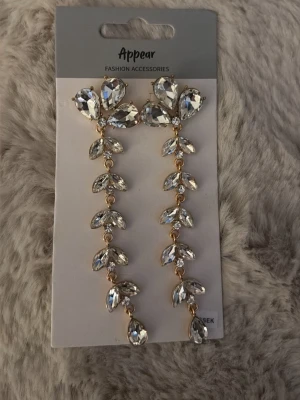 Ingen väst på bilderna - Bilderna visar ett par långa, glittriga örhängen med stora, klara stenar i droppform och guldfärgade detaljer från märket Appear. Ingen väst syns på bilderna.