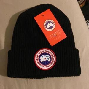 Svart ribbstickad mössa Canada Goose - Svart ribbstickad mössa från Canada Goose med klassisk rund logotyp-patch framtill. Mössan är i tjockt, mjukt material och har uppvikt kant för extra värme. Perfekt för kalla dagar och snygg till streetwear-stilen.