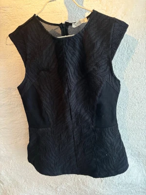 Svart strukturerad ärmlös topp - Säljer en svart ärmlös topp från Zara med snyggt strukturerat tyg i fiskbensmönster och meshdetaljer upptill. Toppen har en figurnära passform och dragkedja i ryggen. 