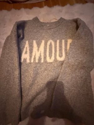 Grå stickad tröja med text från Kappahl - Mysig grå stickad tröja från Kappahl med vit text 'AMOUR' framtill. Tröjan har rund halsringning och långa ärmar, perfekt för kyliga dagar. Enkel och stilren design som passar till det mesta.