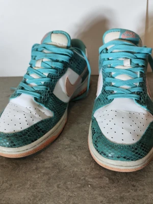 Nike Dunk Low Teal Snakeskin Ormskinn Turquoise EU 42 - Nike Dunk Low “Teal Snakeskin” (DR8577-300). Snygg colorway med turkost ormskinnspräglat material, vit läderbas och peach-färgad swoosh samt sula. Släppt 2022. De är använda men i bra skick. Normalt slitage på yttersula, inga hål eller sprickor. Ormskinnsmaterialet är fint bevarat. Se bilder för exakt skick. Storlek EU 42 (US 8.5). 100% äkta Nike. Mindre creasing på toebox förekommer. Skriv om det är nåt du undrar eller vill diskutera