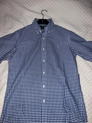 Blårutig skjorta från Ralph Lauren - Säljer en klassisk blårutig skjorta från Ralph Lauren i custom fit. Skjortan har button-down krage, vita knappar och den ikoniska broderade loggan på bröstet. Tillverkad i mjuk bomull och perfekt för dig som gillar stilrena plagg.