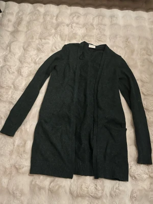 Mörkgrön lång kofta från Vila - Lång, mörkgrön kofta från Vila i mjukt stickat material. Koftan har långa ärmar och praktiska fickor framtill. Perfekt att slänga över en t-shirt eller topp för extra lager. Enkel och stilren design som funkar till det mesta.