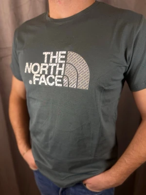 Grå t-shirt från The North Face - Snygg grå t-shirt från The North Face med stort vitt logotryck på bröstet. Klassisk rund hals och korta ärmar. Tillverkad i mjuk bomull som känns skön mot huden. Perfekt för dig som gillar streetwear och vill ha en enkel men stilren look.