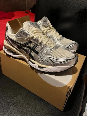 Asics Gel-Kayano 14 sneakers silver/vit - Säljer ett par Asics Gel-Kayano 14 sneakers i silver och vitt med svarta detaljer. Skorna har mesh-överdel för bra andningsförmåga, syntetiska paneler och klassisk snörning. Sulan har tydlig gel-dämpning och retroinspirerad design. Perfekta för dig som gillar sportig stil.