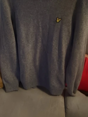 Grå stickad tröja från Lyle & Scott - Mysig grå stickad tröja från Lyle & Scott med deras klassiska gula fågel-logga på bröstet. Tröjan har lång ärm och ribbade muddar vid ärmslut och nederkant. Perfekt för kyliga dagar och enkel att matcha med jeans eller chinos.